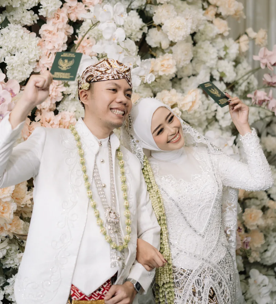 Intan dan Agas dalam busana pengantin di depan rangkaian bunga